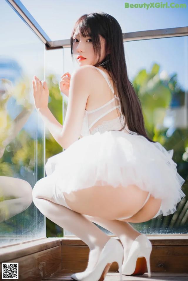 A woman in a white dress and high heels posing by a window.