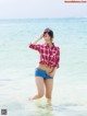 A woman in a red and white plaid shirt and blue shorts standing in the ocean.