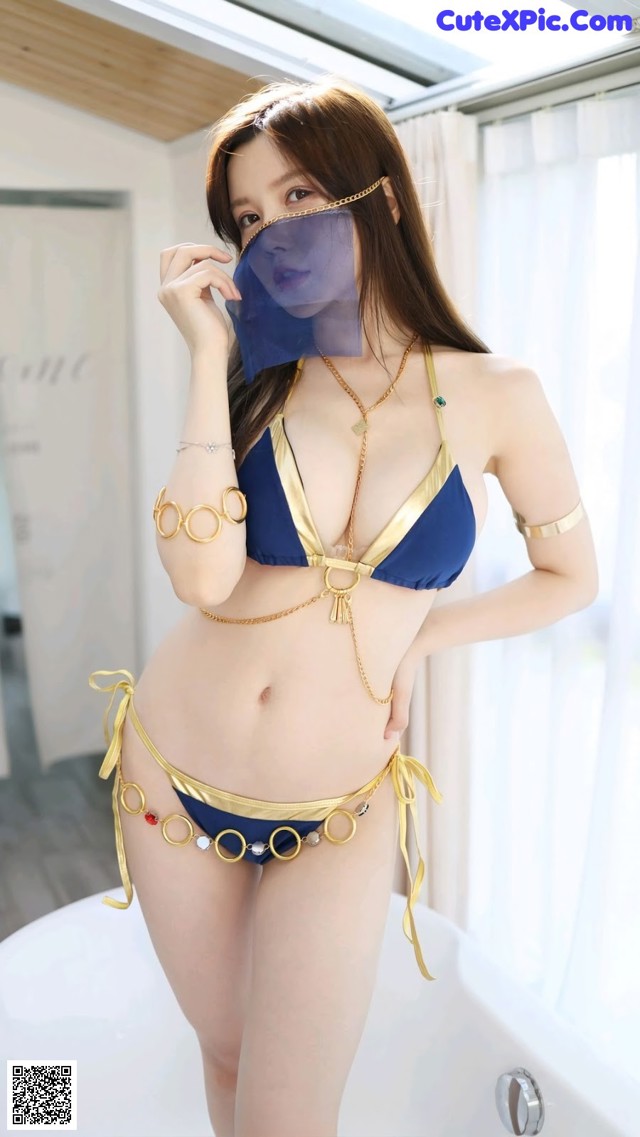 A woman in a blue and gold bikini posing in a bathtub.
