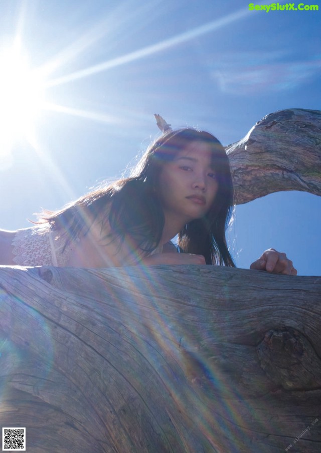 A woman leaning on a tree branch in front of the sun.